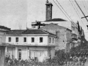 Στιγμιότυπο από την πομπή της κηδείας του δημάρχου Δημητρίου Καραθάνου (18 Μαρτίου 1954). Διακρίνονται τα κτίρια της αριστερής πλευράς της ανηφορικής οδού Βασ. Σοφίας (Παπαναστασίου) και το ρολόι με την ελληνική σημαία μεσίστια.