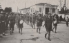 Στιγμιότυπο από την προετοιμασία παρέλασης εθνικής εορτής μιας ομάδας προσκόπων επί της οδού Ηπείρου, μπροστά στην εκκλησία του Αγίου Νικολάου. Δεξιά η κατοικία της οικογένειας Πρωτοσύγκελου. 1948. Αρχείο Φωτοθήκης Λάρισας