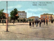 Τα κτίρια της νοτιοδυτικής πλευράς της Κεντρικής πλατείας (Θέμιδος). Χρωμολιθόγραφο επιστολικό δελτάριο του Κωνσταντίνου Παρασκευόπουλου από τον Βόλο. Περίπου 1920.