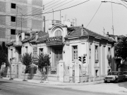 Τα &quot;Δίδυμα σπίτια&quot; της οδού 31ης Αυγούστου. Πίσω από το γωνιακό σπίτι διακρίνεται μέρος της σοφίτας από το τρίτο όμοιο σπίτι, ιδιοκτησίας του Ηλία Τσιτσιά, το οποίο κατεδαφίσθηκε το 1994. Αριστερά η πολυκατοικία που κατασκευάσθηκε δίπλα από το σπίτι του Σταύρου Οικονόμου βρίσκεται ακόμη στο στάδιο κατασκευής. Φωτογραφία προ του 1990. Αρχείο Φωτοθήκης Λάρισας