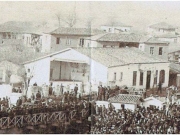 Το θέατρο «Απόλλων» αριστερά της εξόδου της γέφυρας του Πηνειού προς τον Πέρα Μαχαλά.  Λεπτομέρεια φωτογραφίας από την εορτή των Θεοφανείων του 1905. Αρχείο Γιάννη Ρούσκα