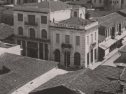 Το Φαρμακείο Καραθάνου στη γωνία των οδών Βενιζέλου και Παπαναστασίου, απέναντι από το κτίριο της Τραπέζης Λαρίσης. Το βέλος υποδεικνύει τη στέγη του Φαρμακείου. Λεπτομέρεια από επιστολικό δελτάριο του 1935 περίπου. Συλλογή Φωτοθήκης Λάρισας.